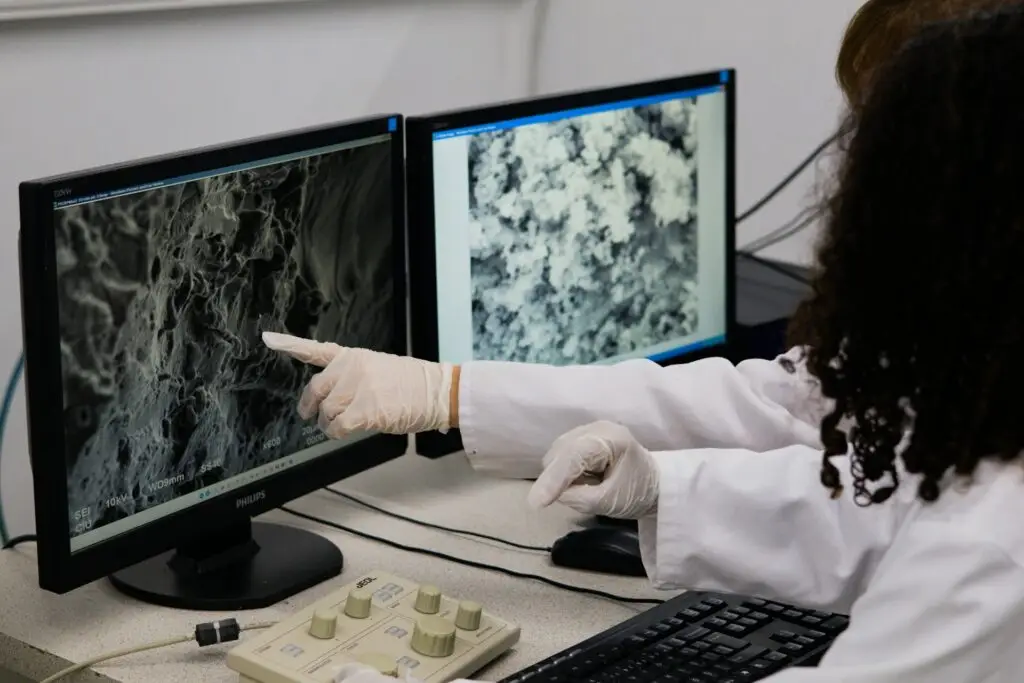 Investigadora con bata y guantes señala imágenes microscópicas en dos pantallas